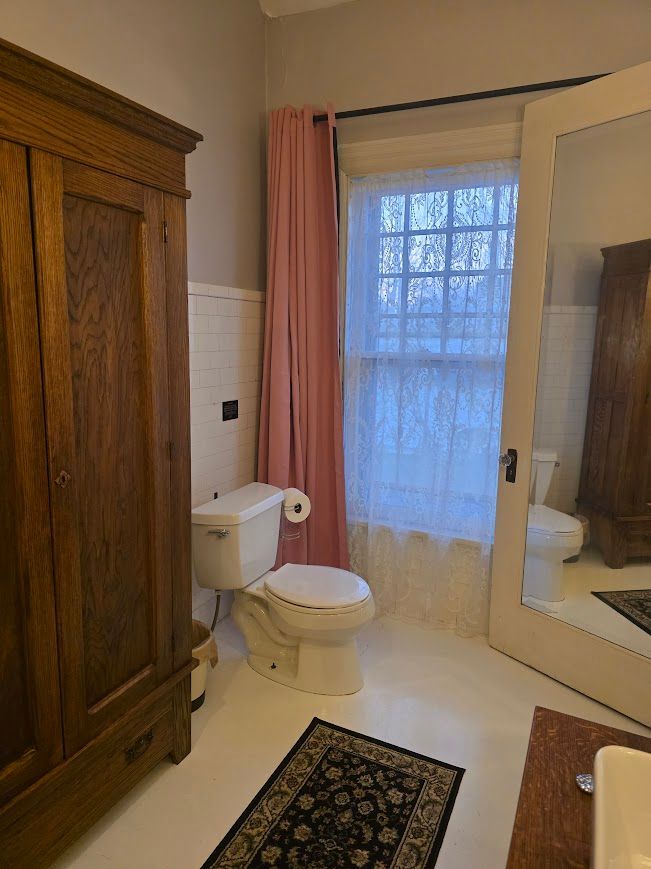 Bathroom with white toilet, pink curtain, vintage wardrobe, and window.