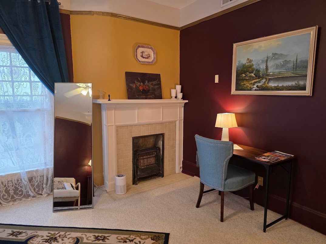 Cozy room with a desk, fireplace, and mirror. Burgundy and gold walls, painting, and blue chair.