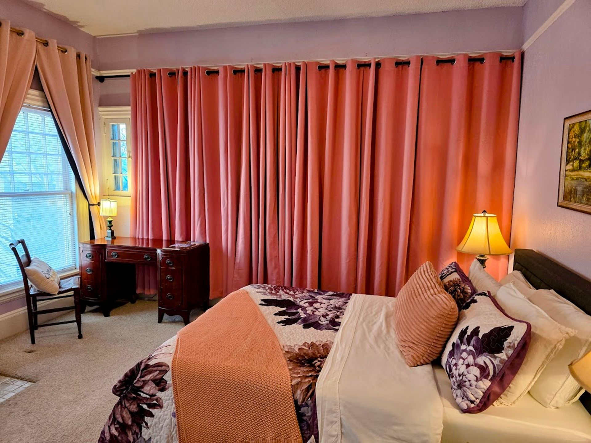 Bedroom with pink curtains, bed with floral comforter, desk, and window.