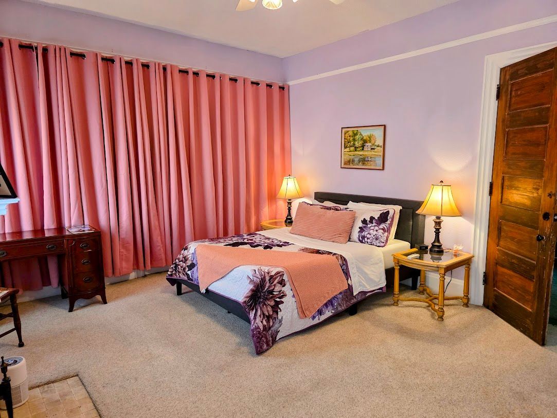Bedroom with pink curtains, bed with purple and pink accents, two lamps, and a wooden door.