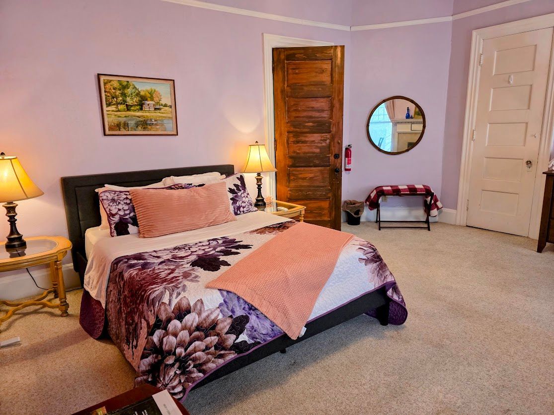 Bedroom with lilac walls, a bed with floral bedding, wooden door, and two lamps.