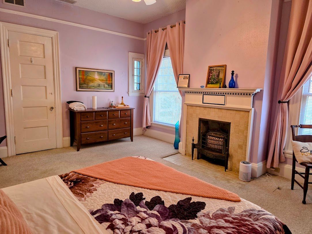 Bedroom with lilac walls, wooden furniture, a fireplace, and a bed with a floral quilt.