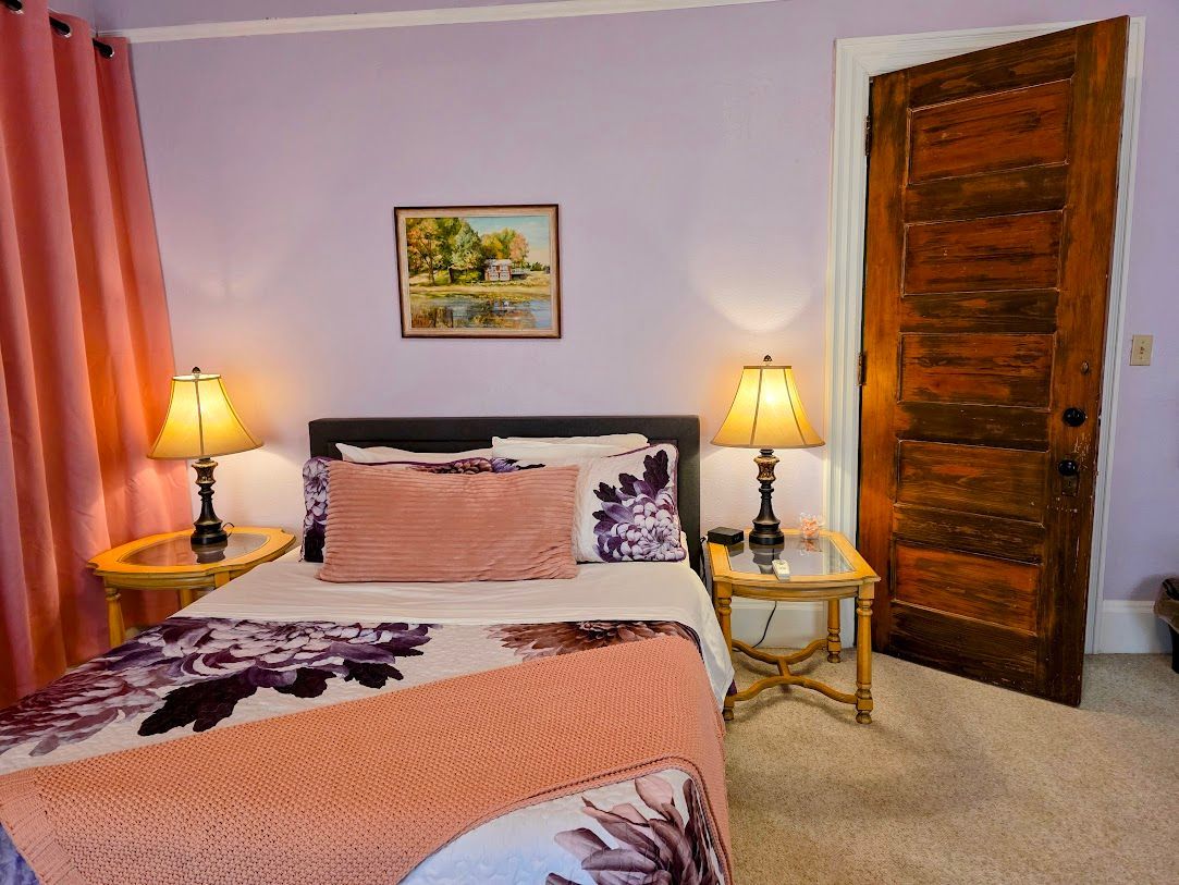 Bedroom with purple walls, bed with floral bedding, pink curtains, bedside tables, and an antique wooden door.