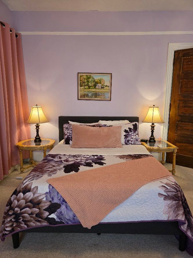Bedroom with purple walls, floral bedding, two lamps on nightstands, and pink curtains.