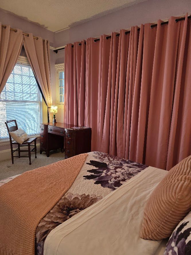 Pink curtains in bedroom; bed, window seat, and small table with lamp.