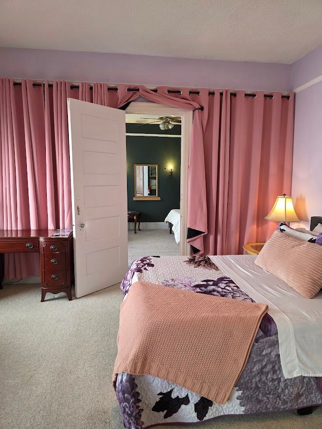 Bedroom with pink curtains, bed, desk, and doorway leading to another room.