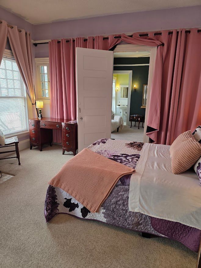 Bedroom with pink curtains, a bed with purple quilt, and an open door to a bathroom.