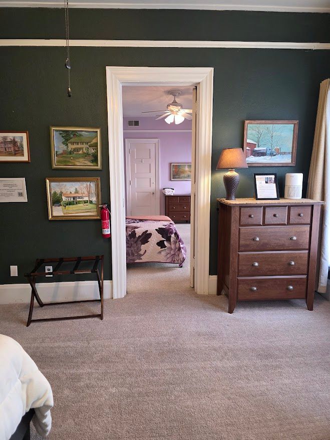 Bedroom doorway looking into a second bedroom. Dark green walls with art, a dresser, and a small bed-side table.