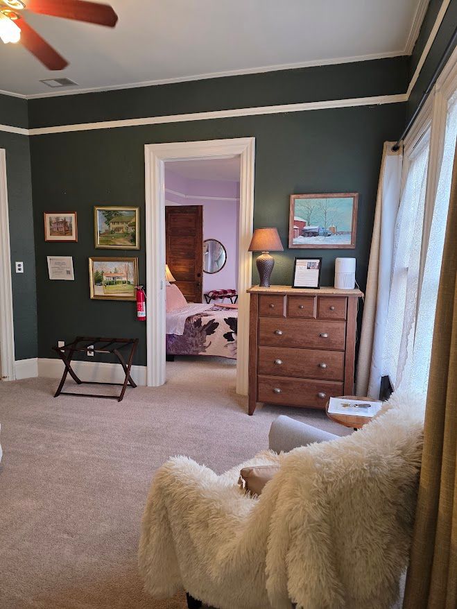 Cozy hotel room with dark green walls, a wooden dresser, and a doorway leading to a bedroom.