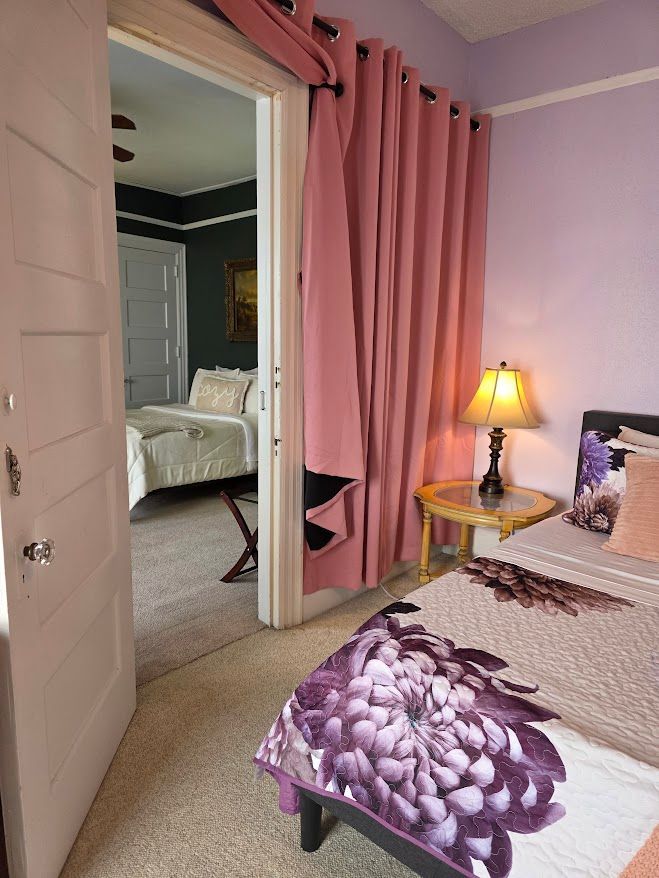A bedroom with a pink curtained doorway opening into another bedroom. Bed and table with lamp in view.