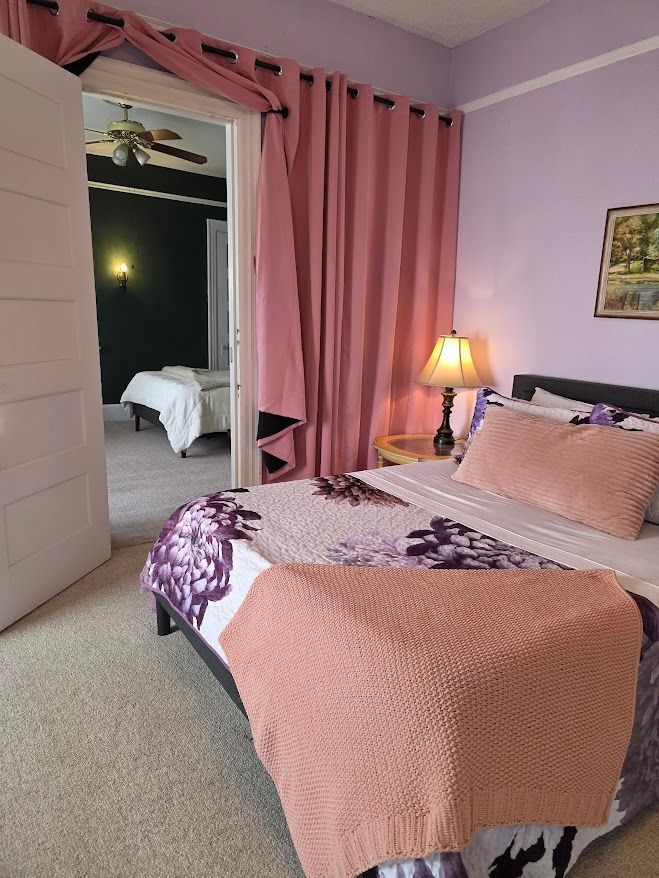 Bedroom with lavender walls, pink curtains, a bed with floral quilt and pink throw, doorway to another room.