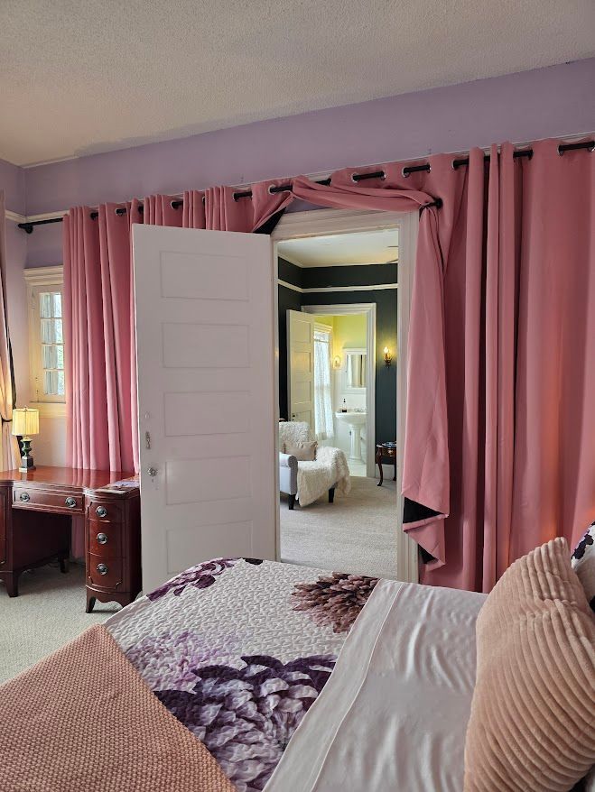 Bedroom with pink curtains, open door to bathroom, desk, and bed with floral bedding.