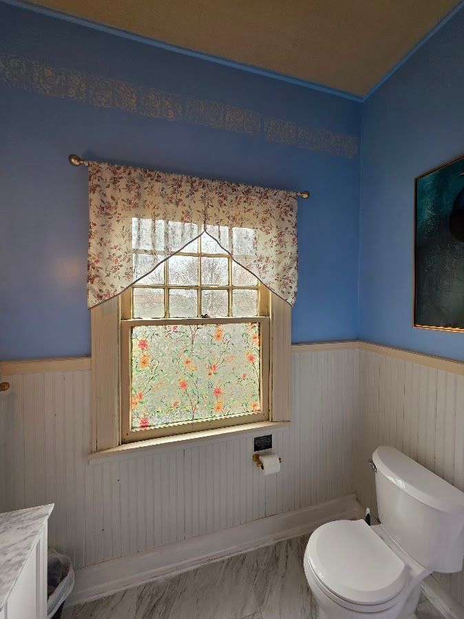 A bathroom corner with blue walls above white wainscoting, a floral-curtained window, and a white toilet.