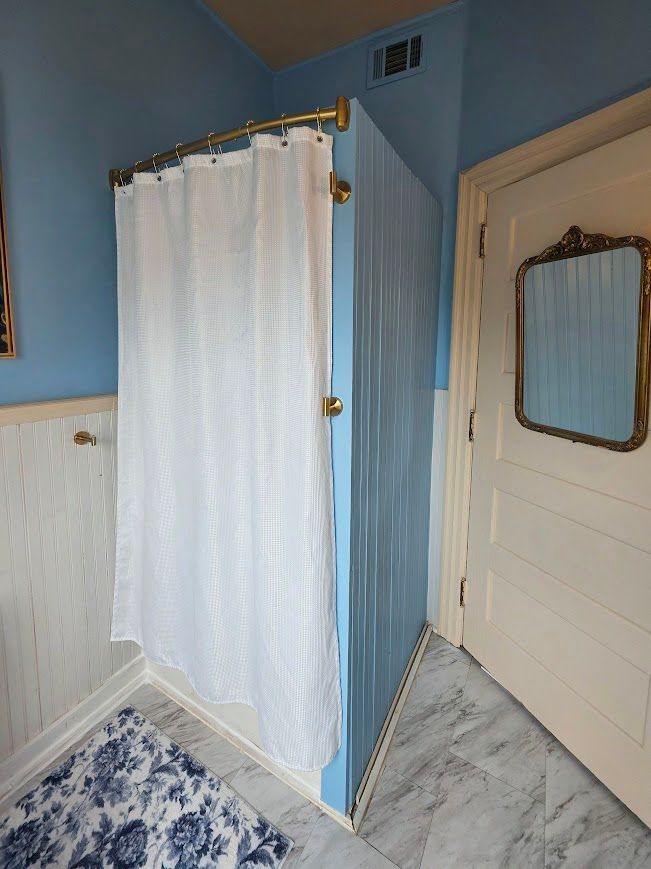 A corner shower with a white curtain, light blue walls, and marble flooring in a bathroom.