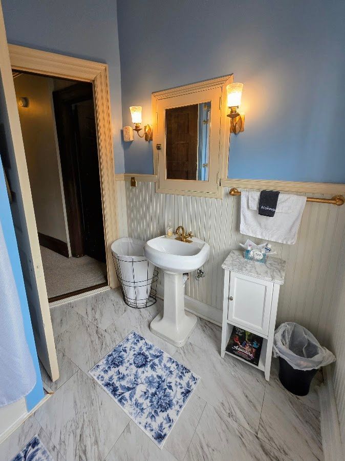 A blue-walled bathroom featuring a pedestal sink, a small white cabinet, a mirror, and a floral patterned rug.