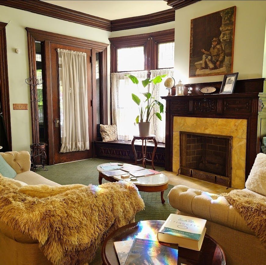 Cozy living room with fireplace, window seat, and couches with fuzzy throws. Sunlight streams across coffee tables.