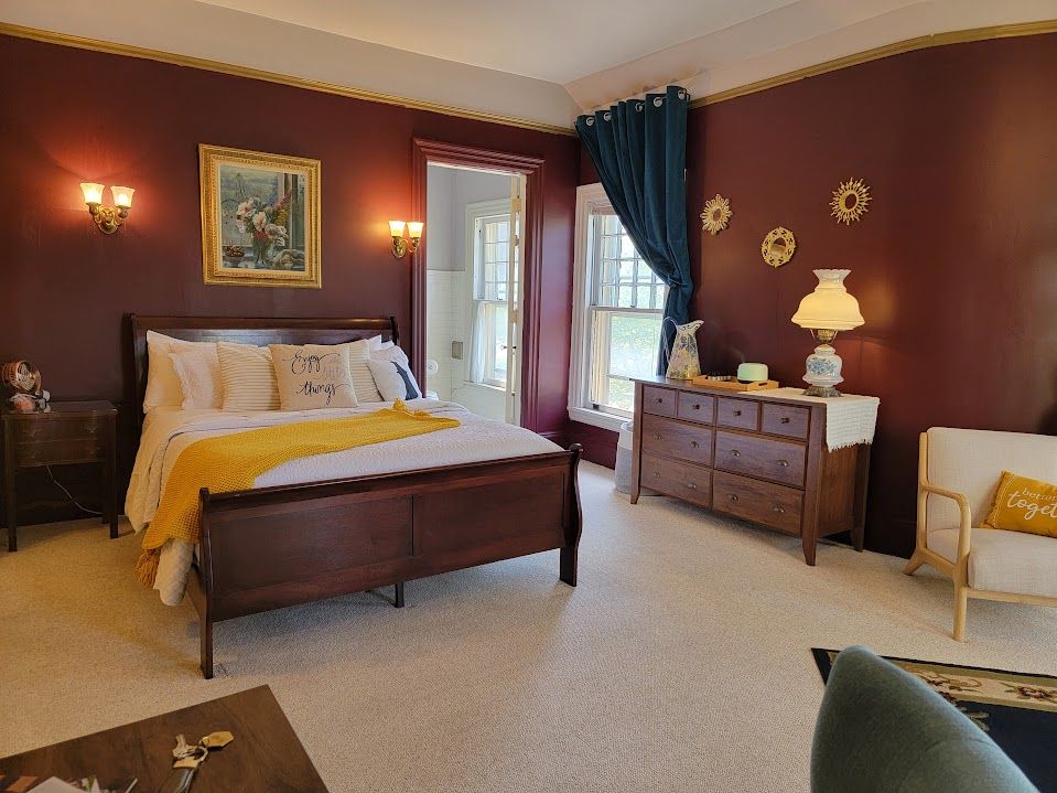 Bedroom with burgundy walls, wooden bed, dresser, and armchair. Window with teal curtain and gold accents.