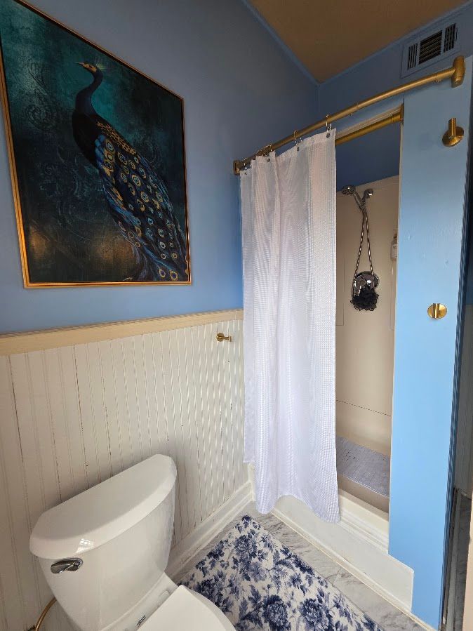 A blue bathroom with a peacock painting, white wainscoting, a toilet, a floral rug, and a shower with a white curtain.