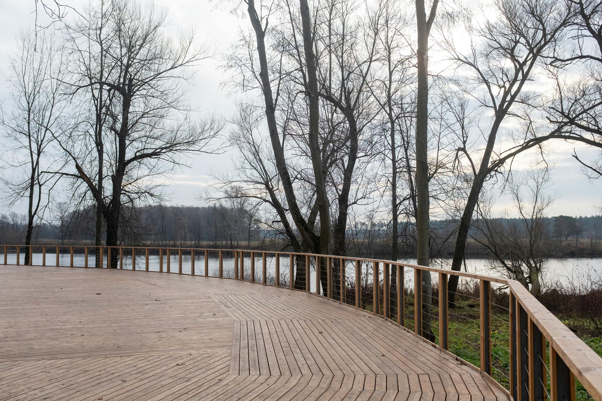 Curved wooden boardwalk with railings beside a calm river and leafless trees on an overcast day