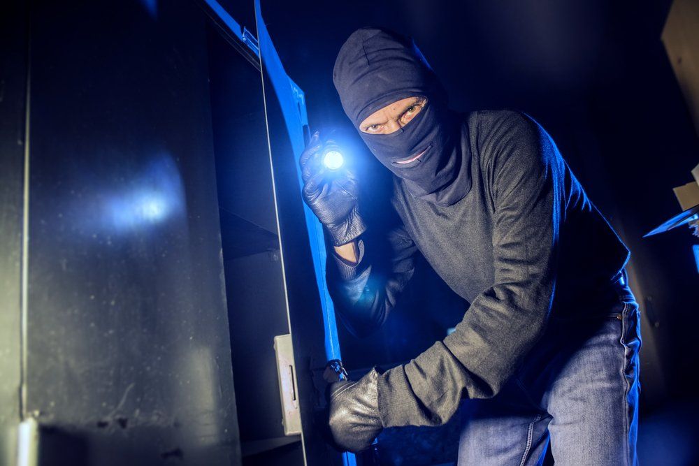 Burglary in progress: masked figure in dark room with flashlight, attempting to open something.