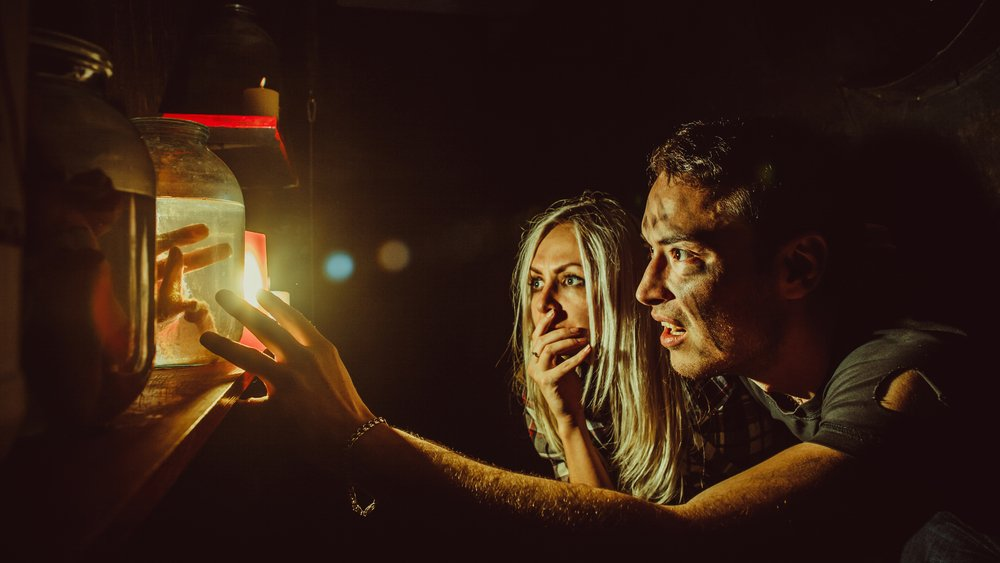 Man and woman gaze at a glowing jar lit by a candle in a dark setting; suspenseful expressions.