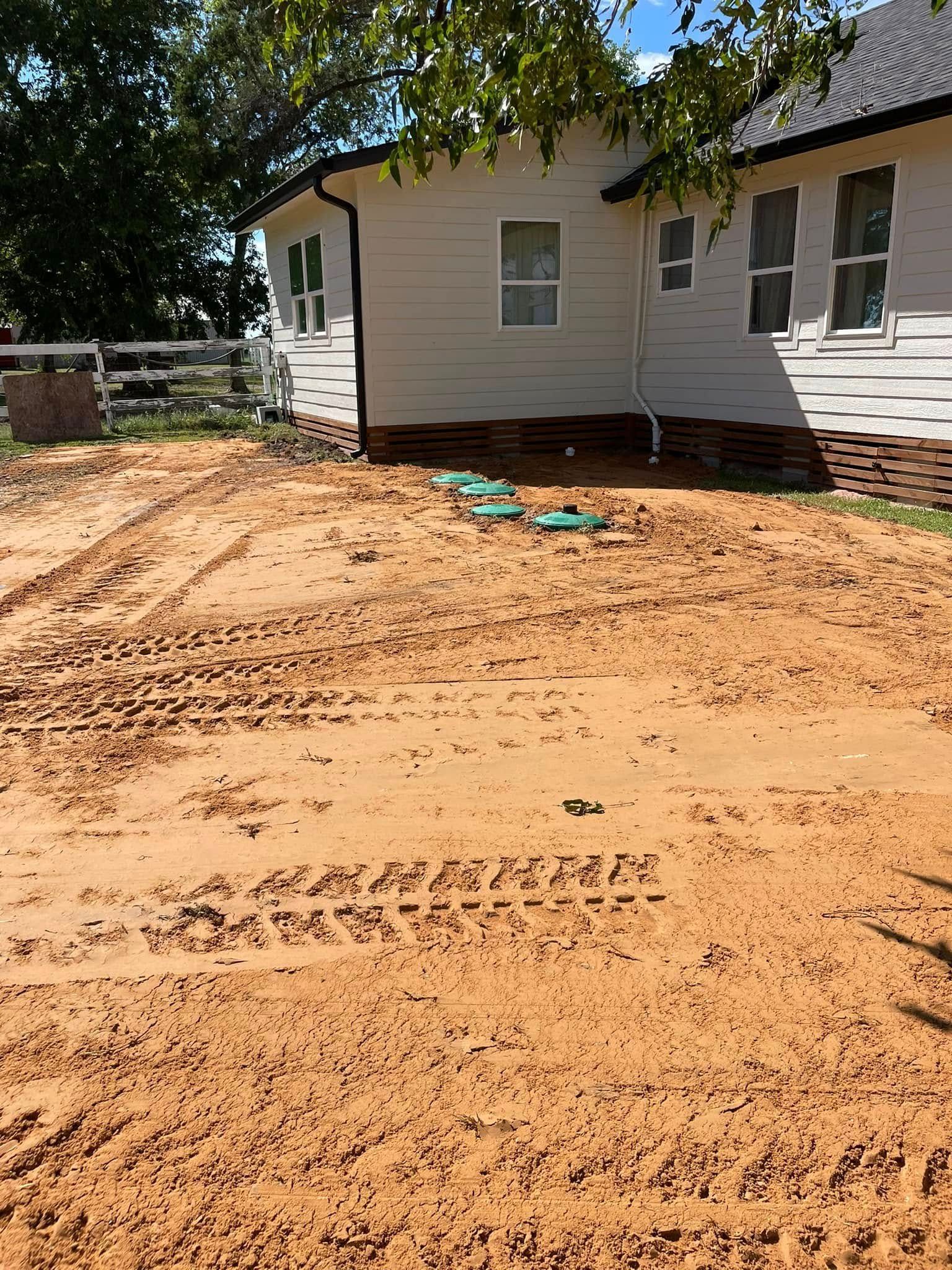 Freshly graded dirt yard beside a white house with green shutters under trees.