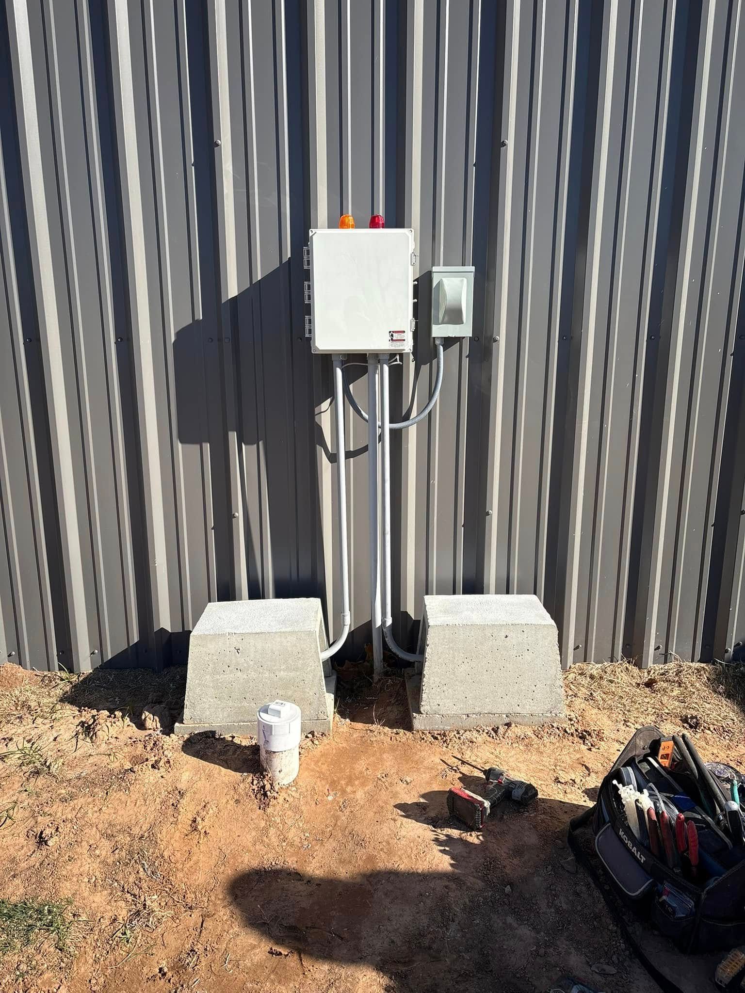 Electrical box and meter mounted on a corrugated metal wall, with concrete blocks and dirt below.