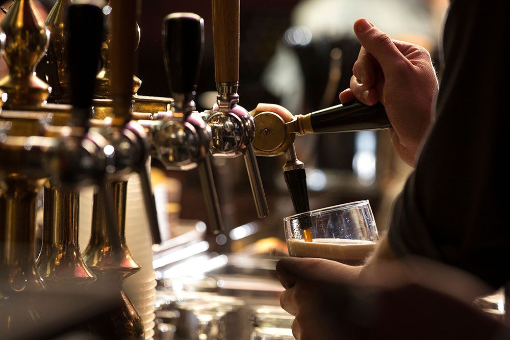 A person pours a glass of dark beer from a brass tap at a bar.