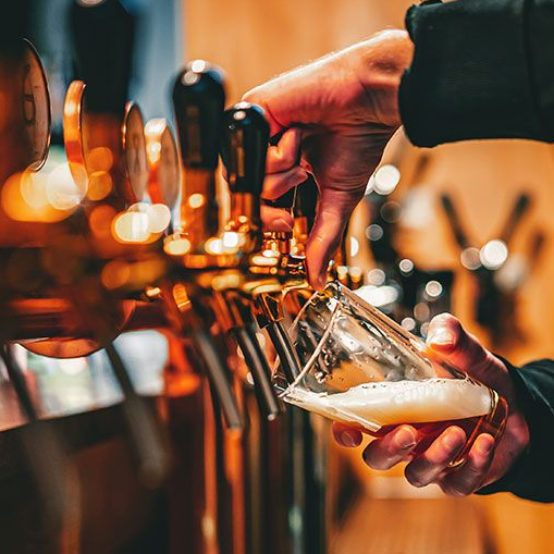A close-up of a hand pouring a beer from a brass tap into a glass, with multiple taps blurred in the foreground.