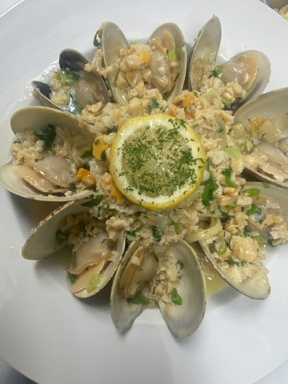 A plate of clams arranged in a circle around a central, herb-topped lemon slice, surrounded by minced clam stuffing.