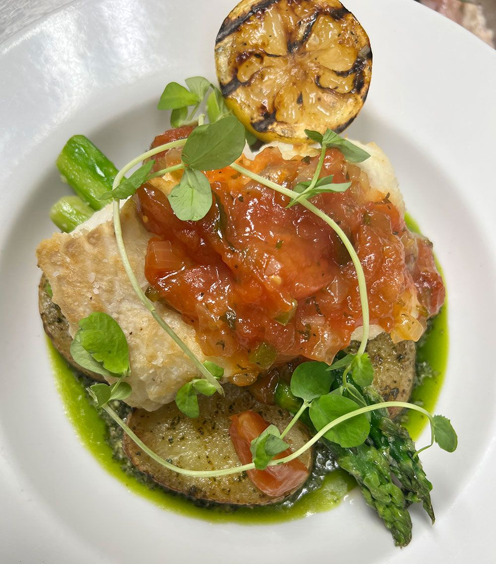 A white bowl containing pan-seared fish topped with tomato relish, asparagus, and grilled lemon, garnished with greens.