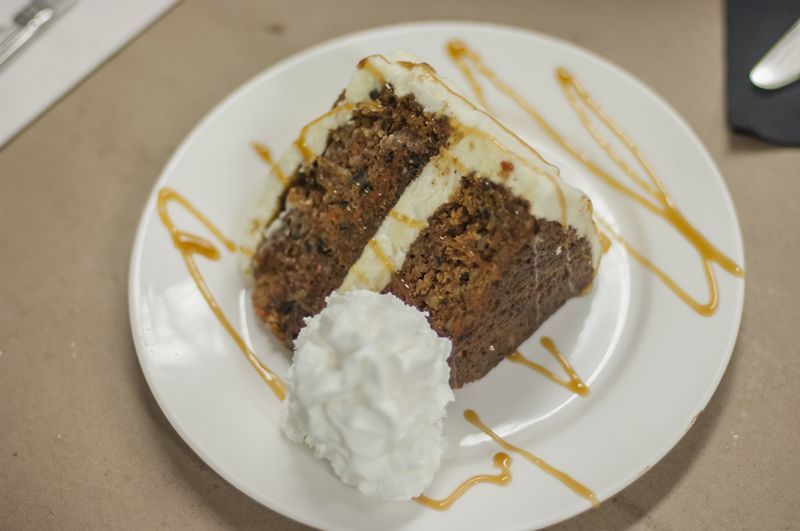 Slice of chocolate cake with frosting, white plate, wine, wooden table.