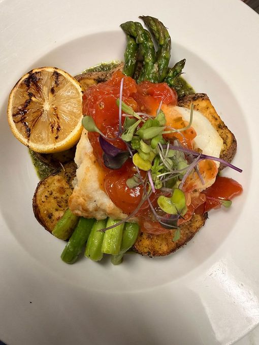 A seared fish fillet topped with tomato relish, served with roasted potatoes, asparagus, and a charred lemon half.