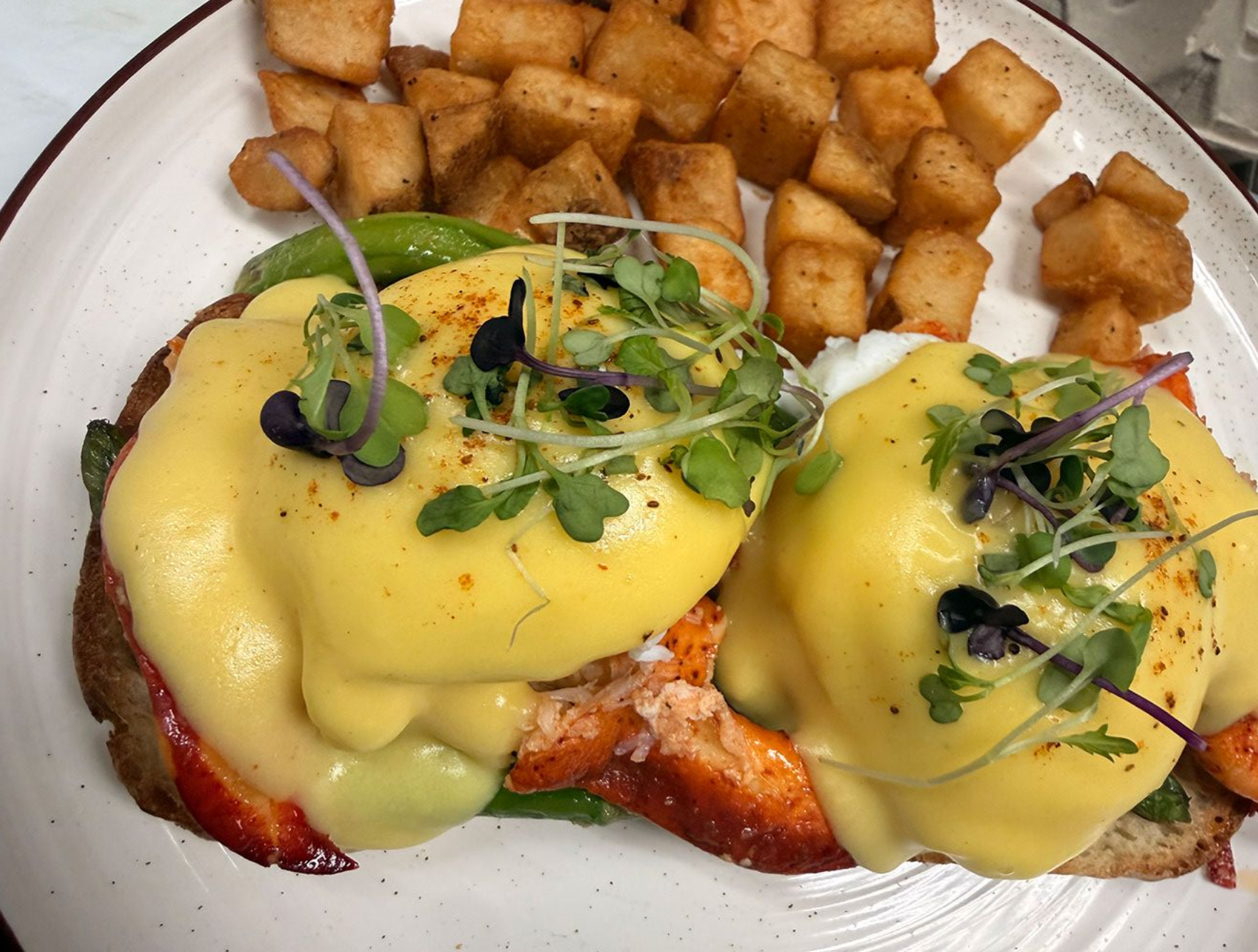 Two lobster eggs Benedict topped with microgreens and hollandaise sauce, served with crispy potato cubes on a white plate.