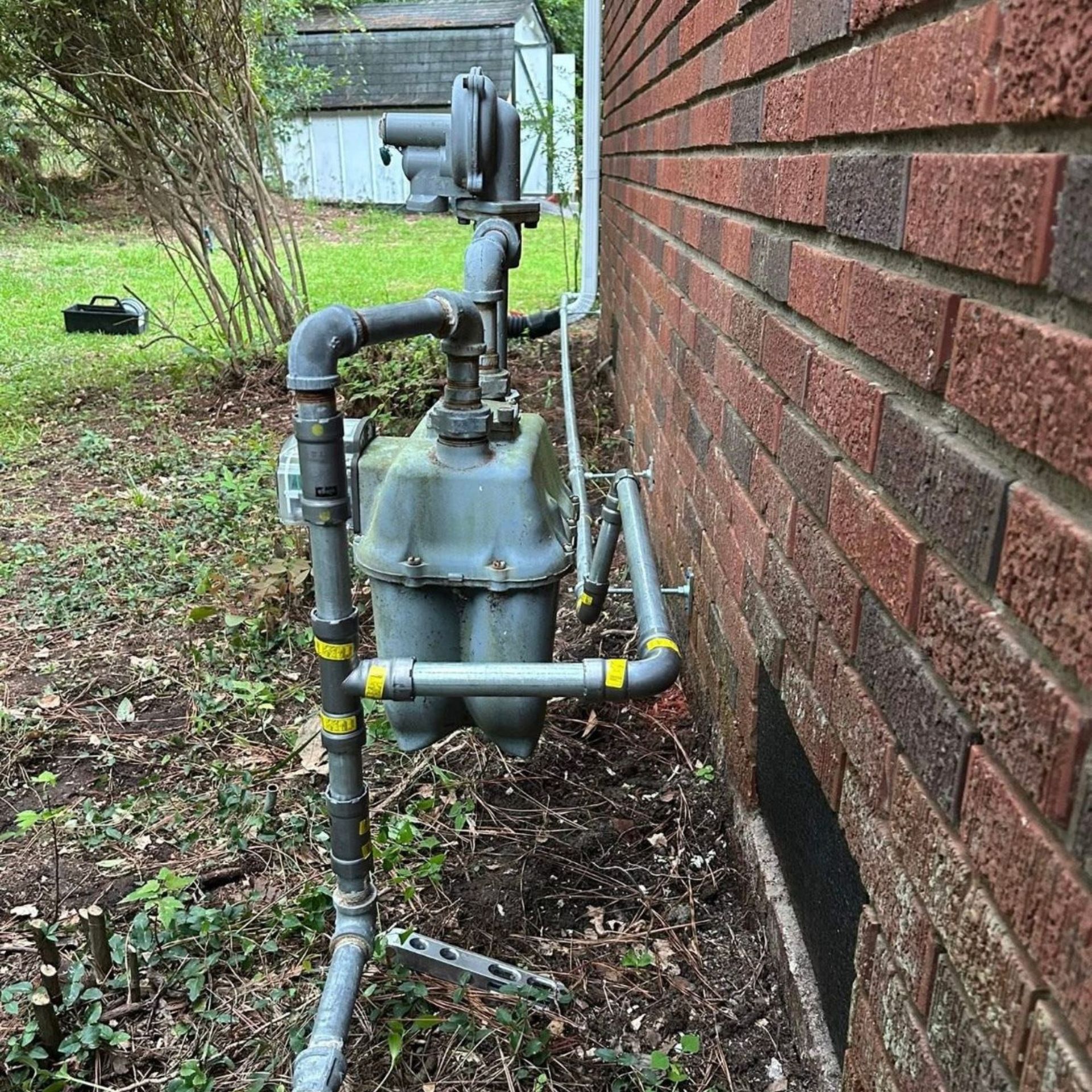 Black River Plumbing & Gas LLC - Gas meter attached to a brick building, with pipes and a regulator. Outdoors, in a yard.