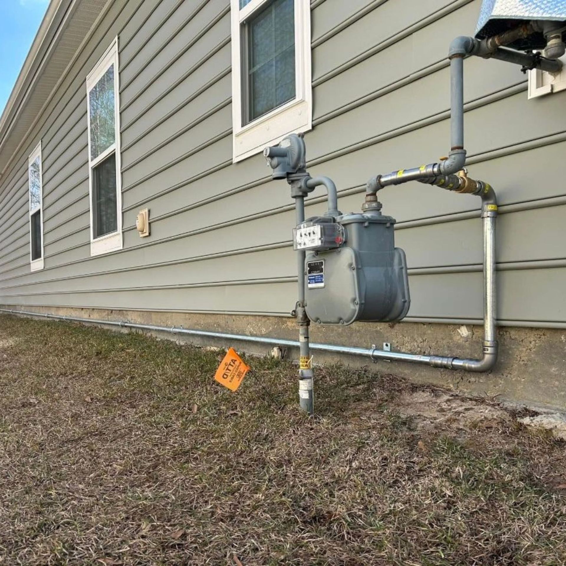 Gas meter and pipes attached to a light gray house exterior on a lawn with a survey flag.