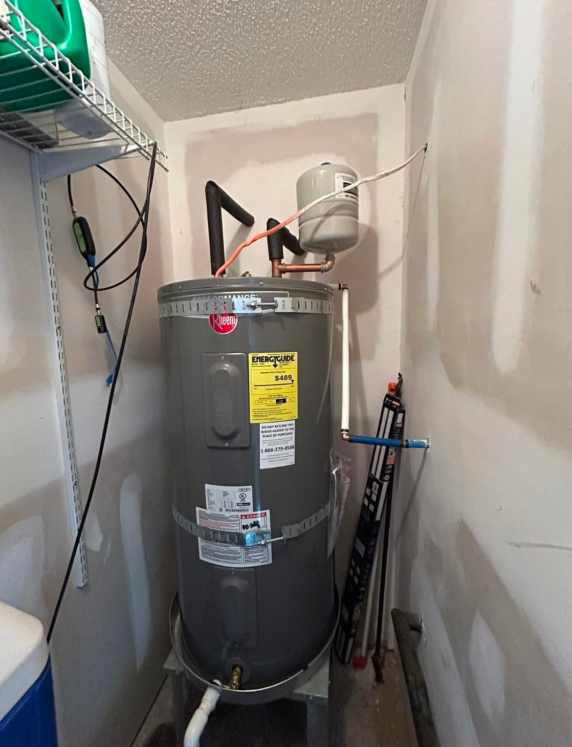 A gray water heater with a small expansion tank in a small room with a shelf and tools.