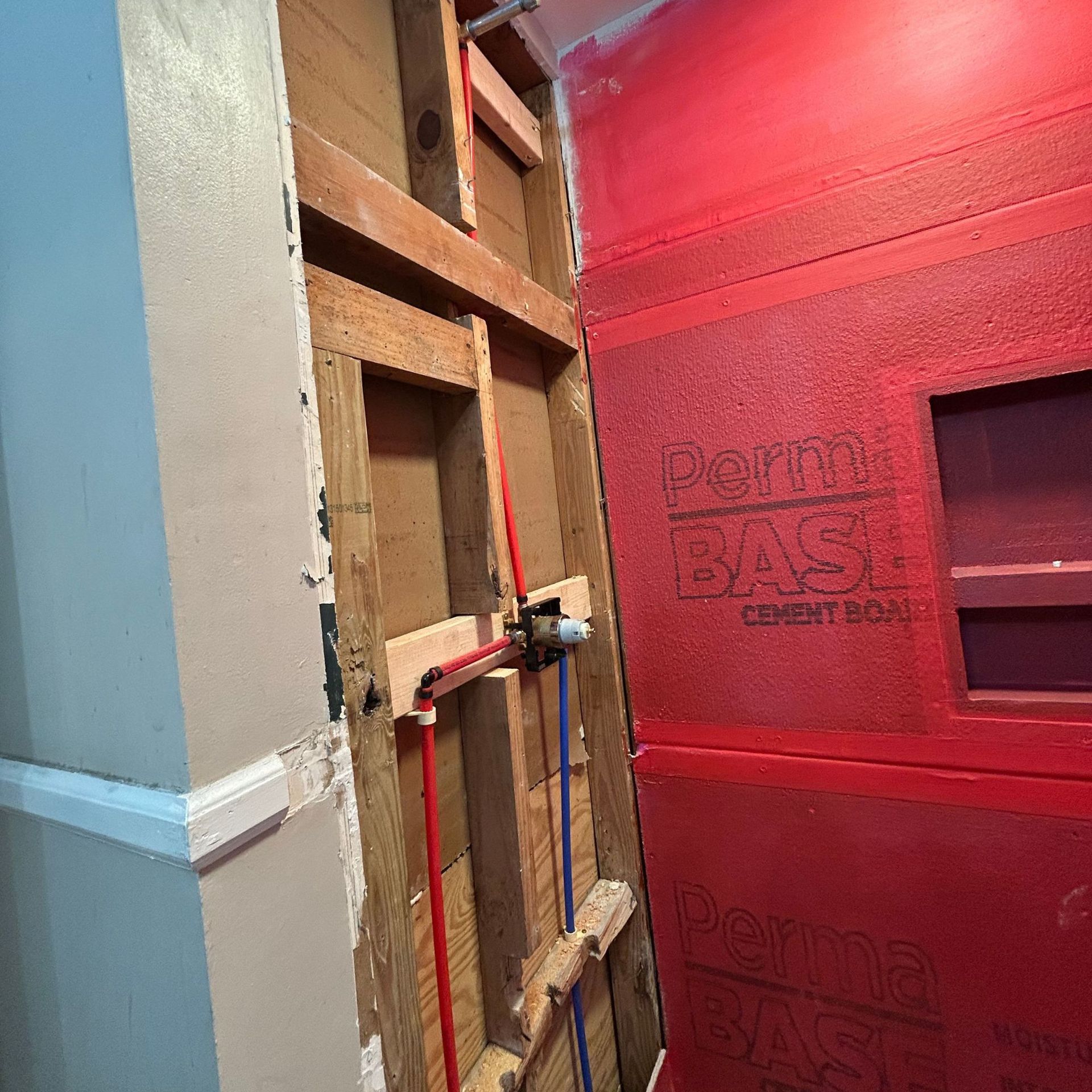 Black River Plumbing & Gas LLC - Bathroom wall under construction, with exposed pipes and cement board. Red and blue pipes.