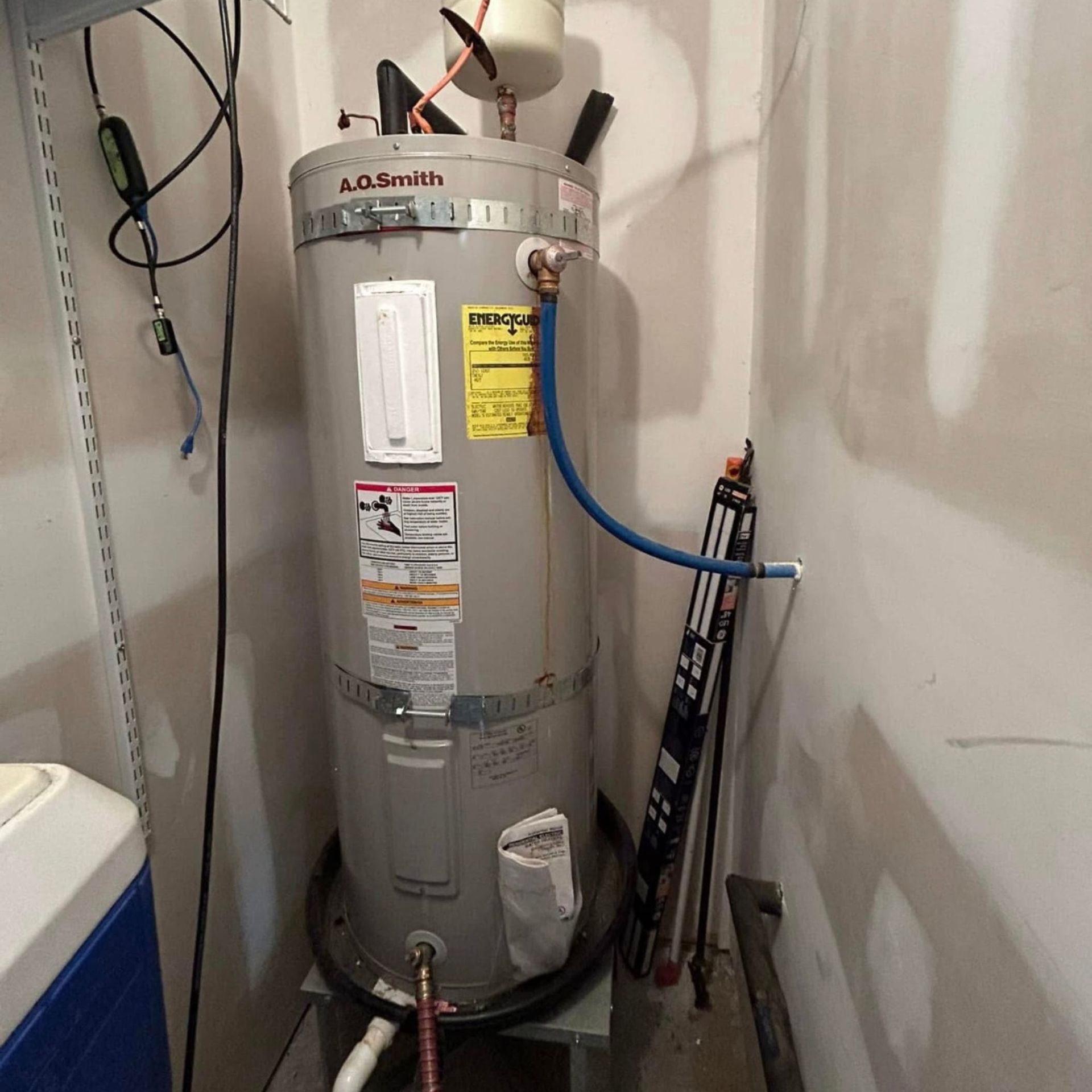 Water heater in a corner. Cylindrical, light gray tank with various pipes, labels, and a blue hose.