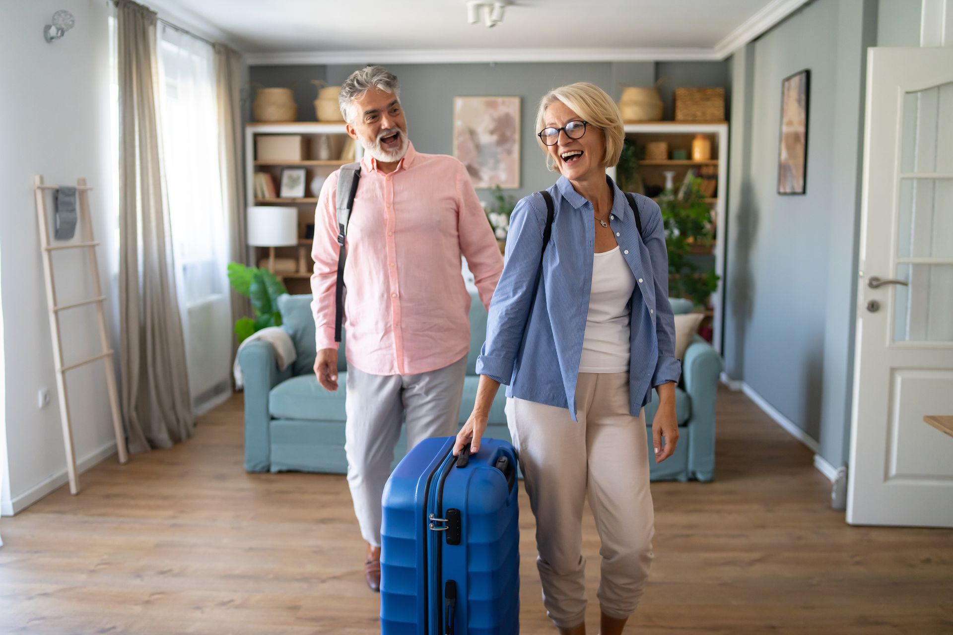 An elderly couple is walking with their luggage in a living room.
