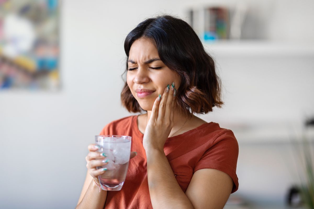 Woman holding ice water, grimacing and touching cheek, possibly from tooth pain.