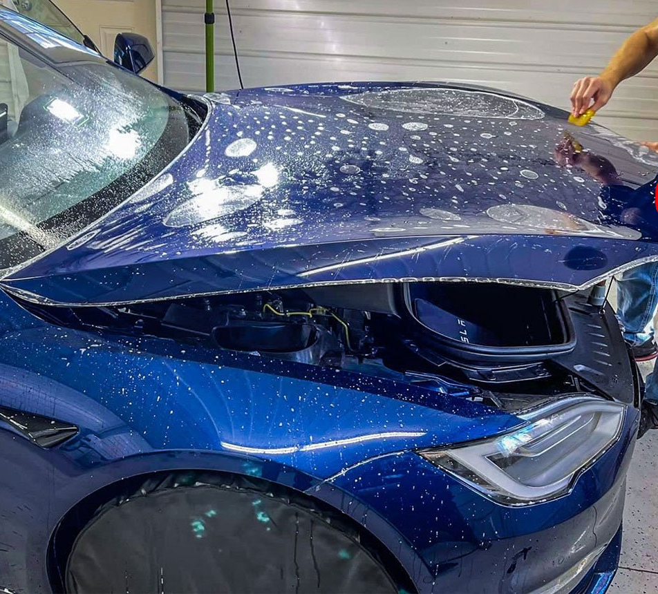 A man is applying a protective film to the hood of a blue car.