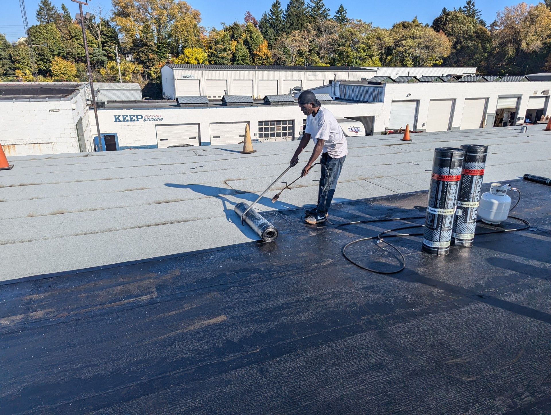 Man on a flat roof using a torch to apply roofing material. Rolls of material and propane tank visible. Sunny day.