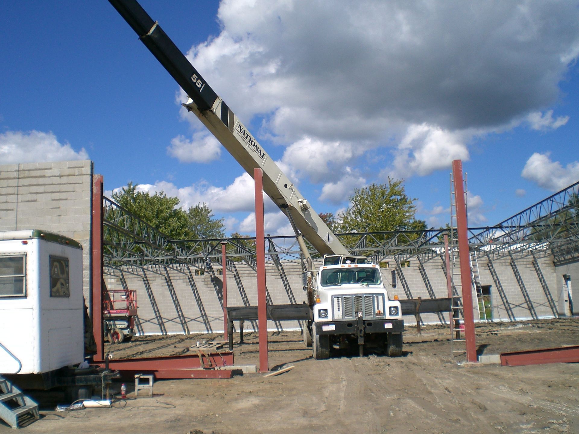 Crane lifting a concrete beam at a construction site; white truck, red columns, blue sky.