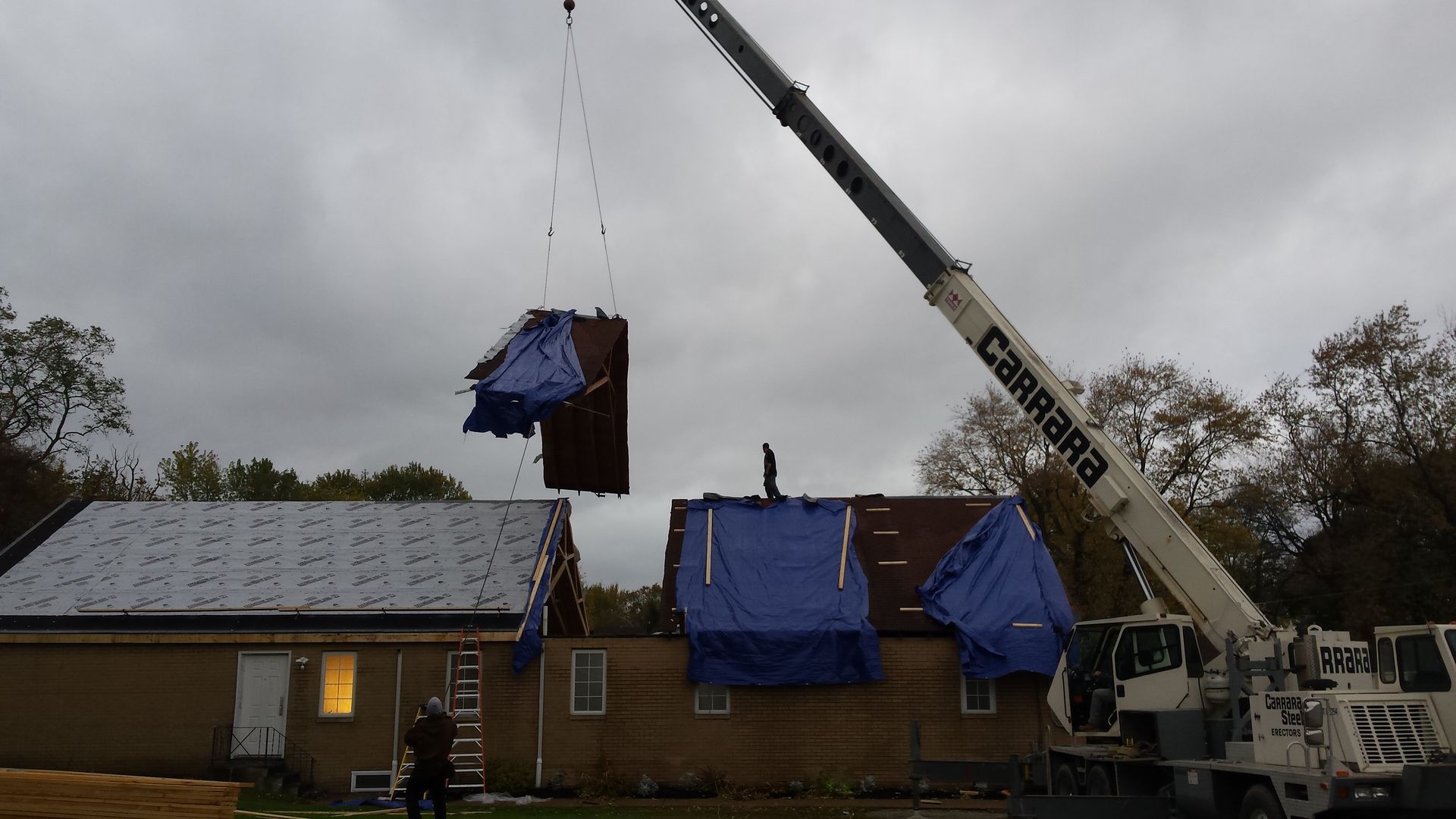 A crane lifting a large roof section off a house covered in blue tarps, under a cloudy sky.