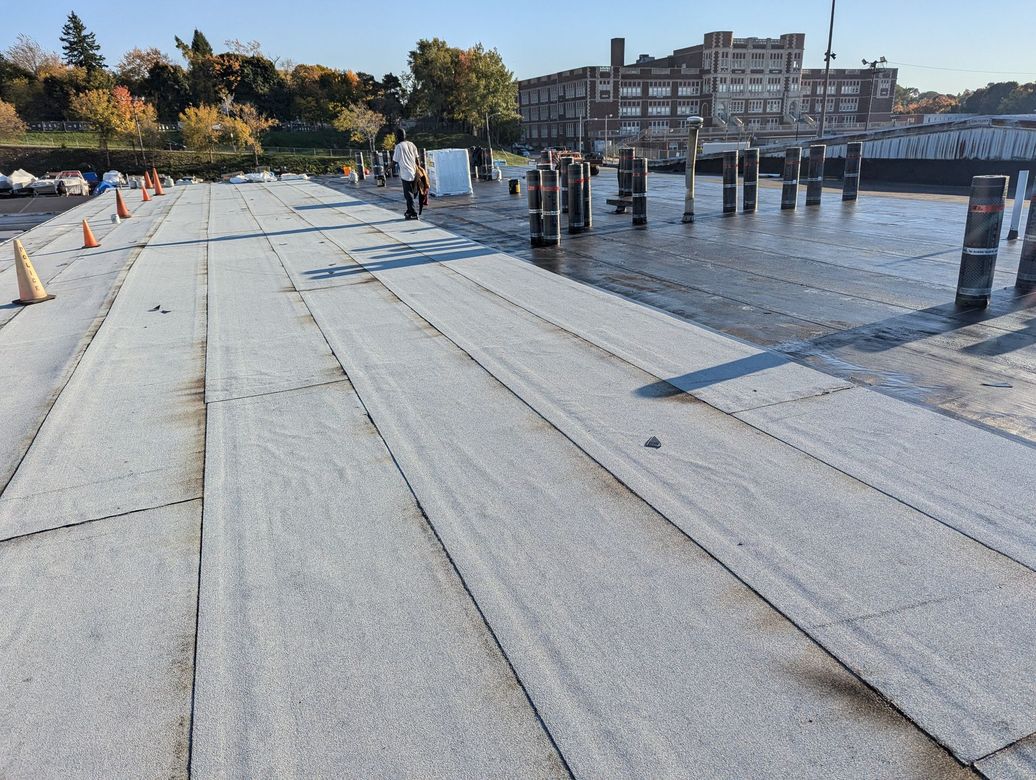 Rooftop being worked on; rolls of roofing material laid out, workers, and a distant brick building.