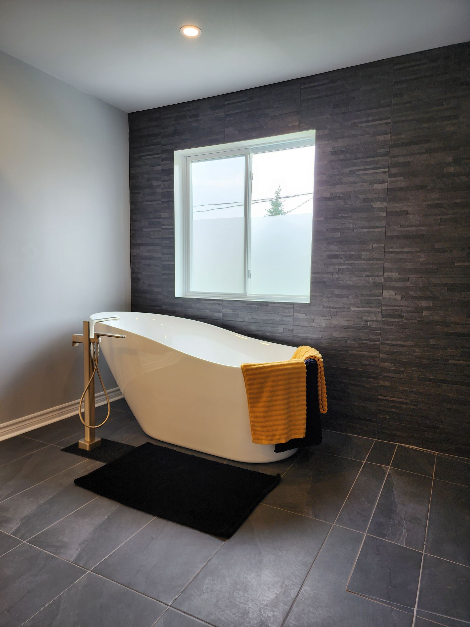 Salle de bain avec baignoire blanche, murs carrelés foncés, fenêtre et tapis de sol noir.