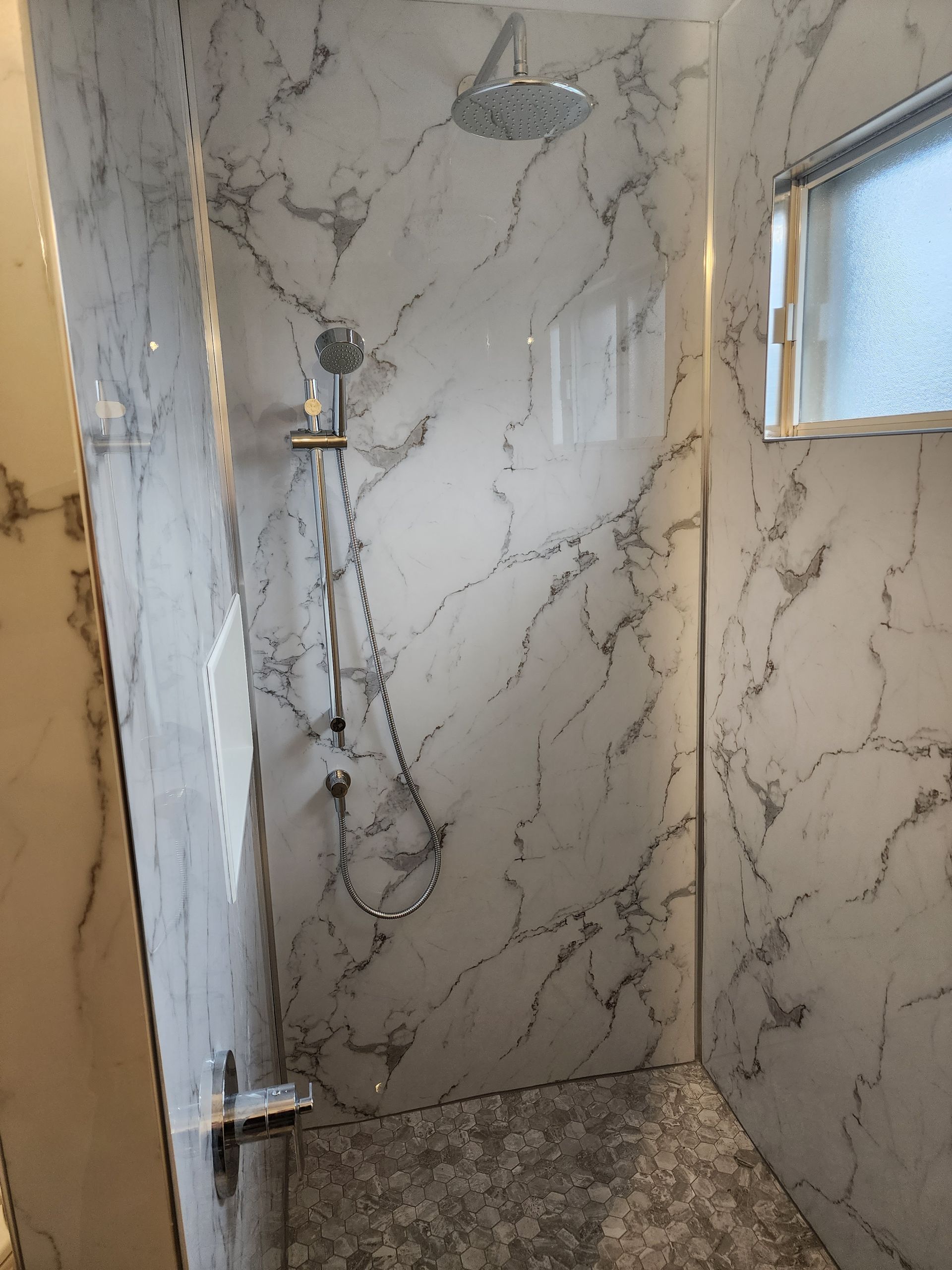 Douche carrelée en marbre avec robinetterie argentée. Une petite fenêtre laisse entrer la lumière.