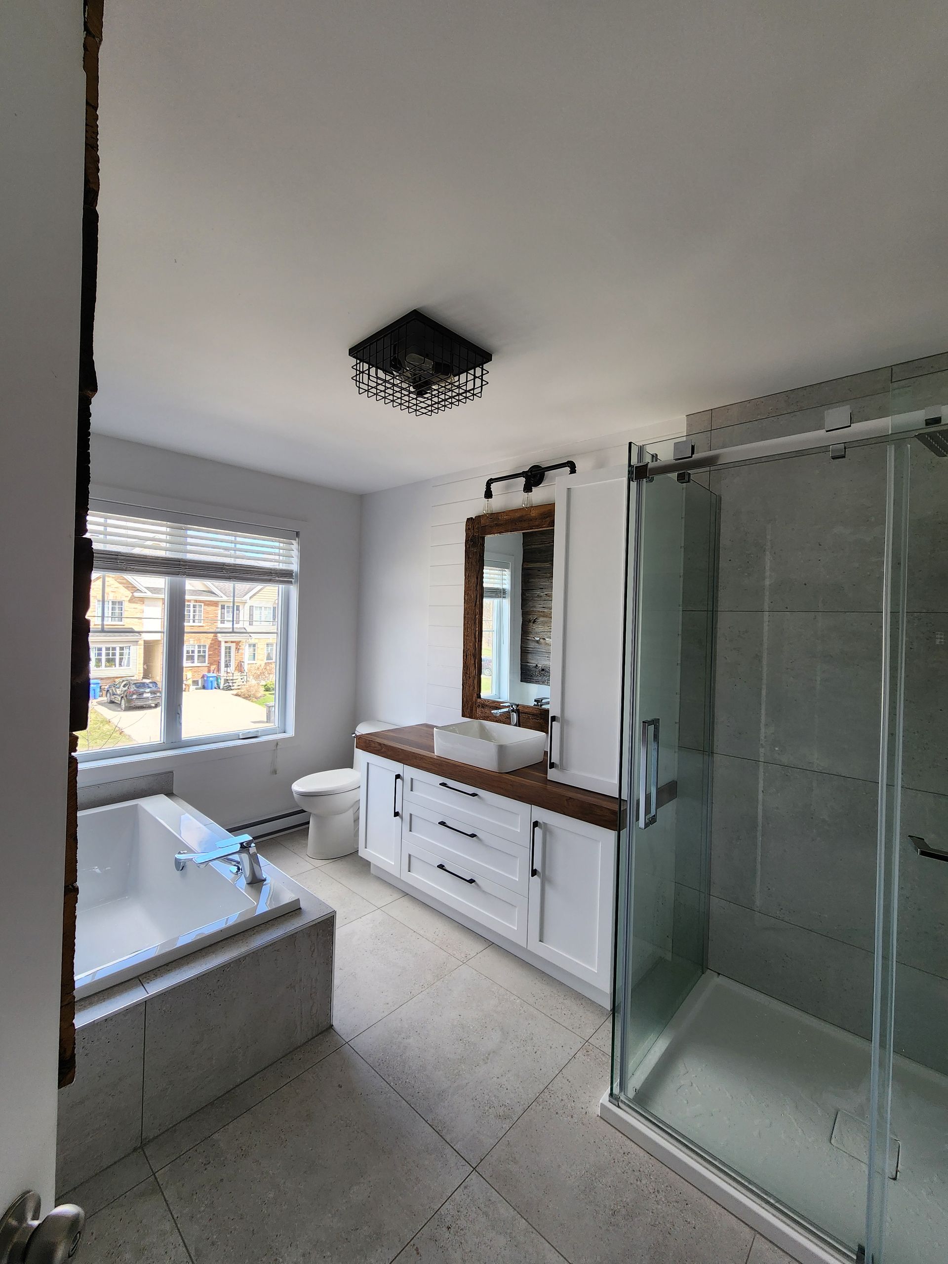 Salle de bain avec vanité blanche, douche à l'italienne, baignoire et fenêtre donnant sur une rue.