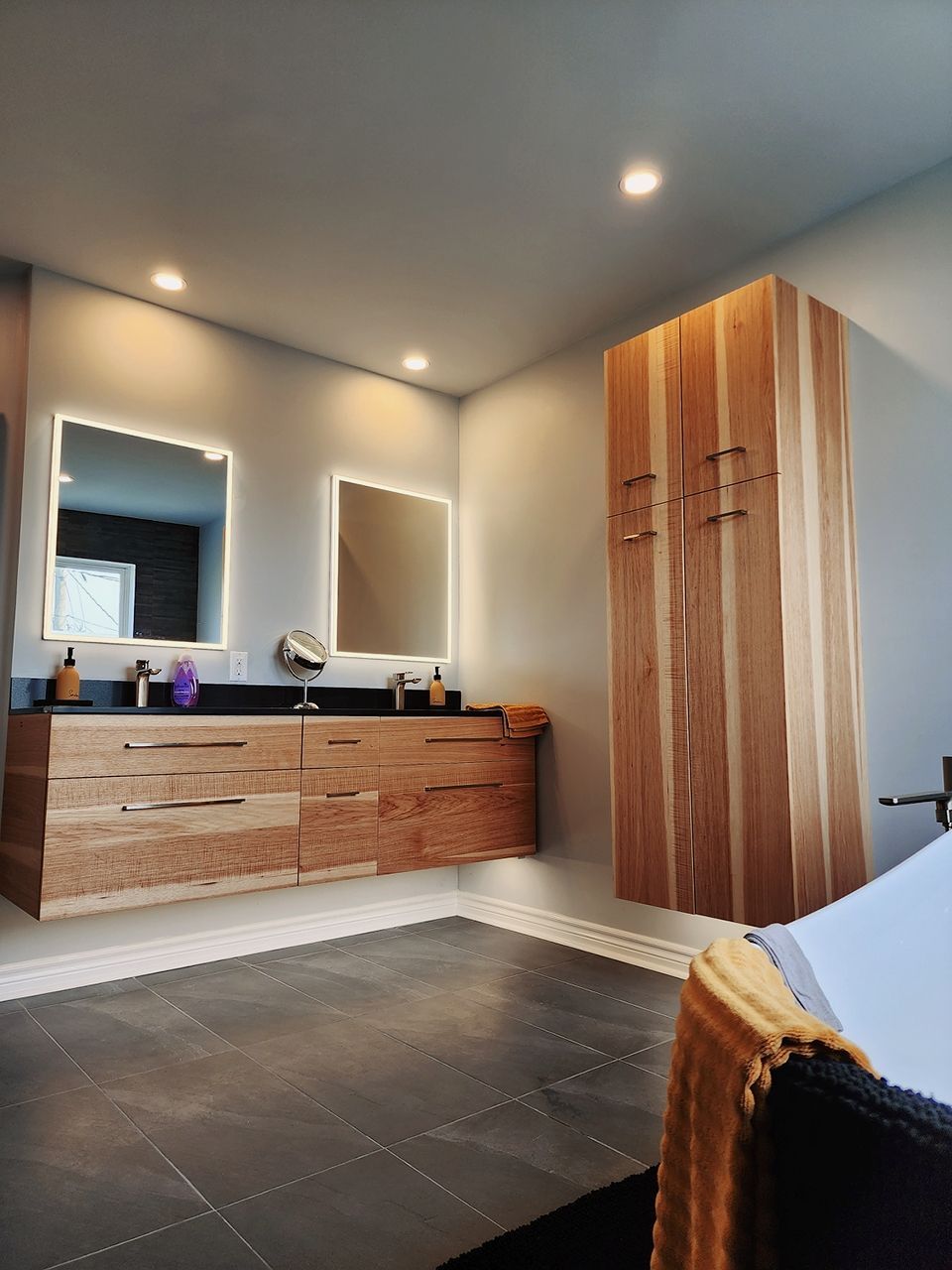 Salle de bain avec vanité en bois, deux miroirs, grande armoire de rangement et sol en carrelage gris.